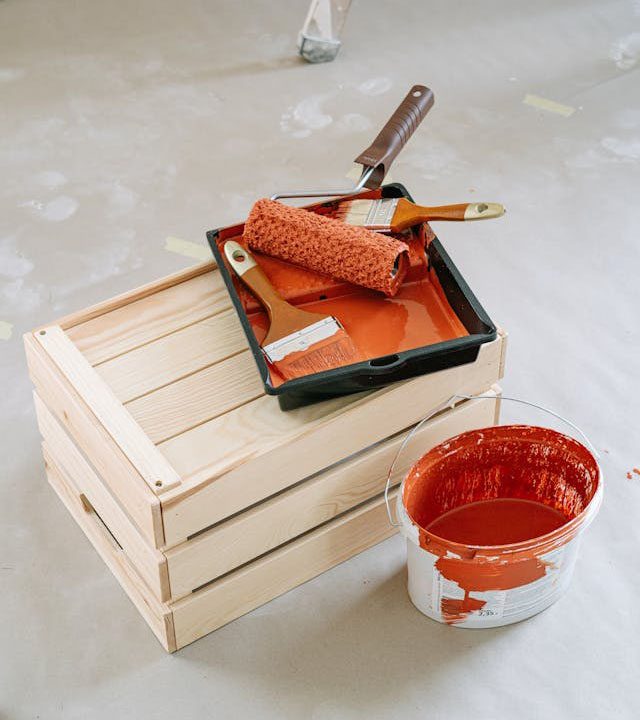 Painting supplies including brushes, a roller, a paint tray with red paint on a wooden crate, and a paint bucket on the floor.