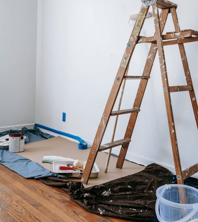 Wooden ladder, paint cans, brushes, roller tray, and drop cloths set up for indoor wall painting in a room with hardwood floors.